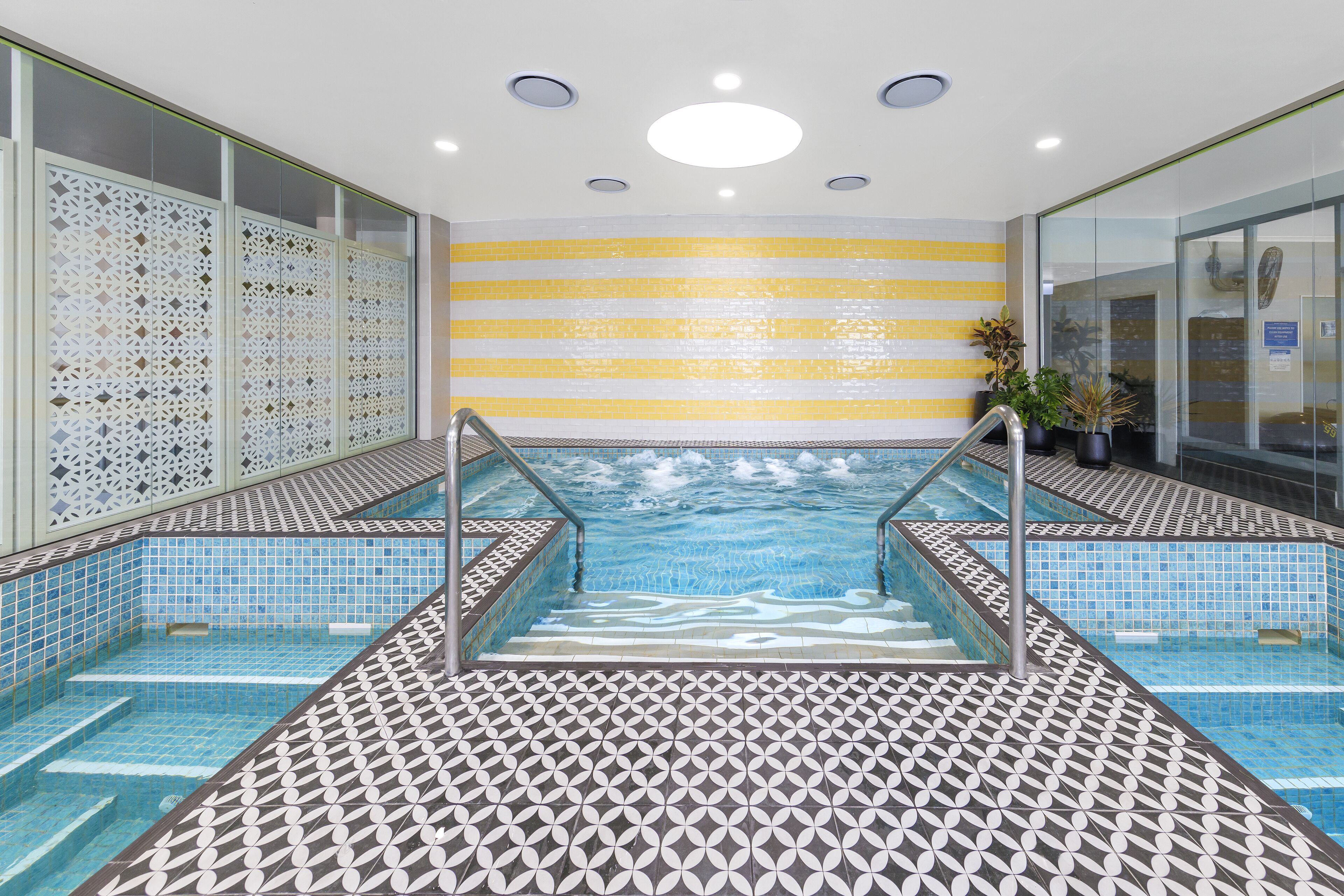 A swimming pool with a geometric tile pattern, a yellow and white striped wall, and a glass partition with a decorative screen.