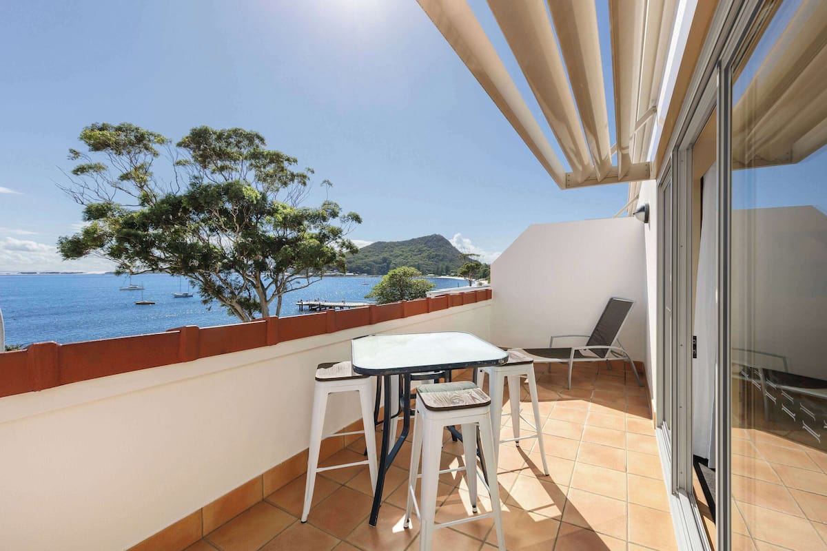 Ein Balkon mit Tisch und Stühlen, der auf ein Gewässer mit Booten und ein entfernteres Gebirge blickt.
