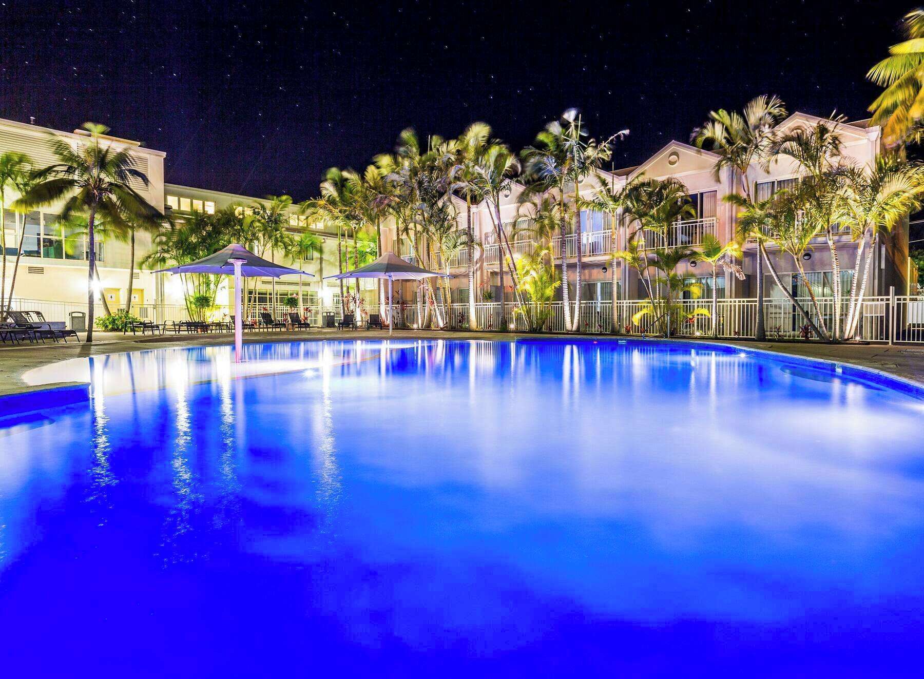 Un área de piscina nocturna con una piscina azul cristalina, palmeras y una hilera de edificios con balcones.