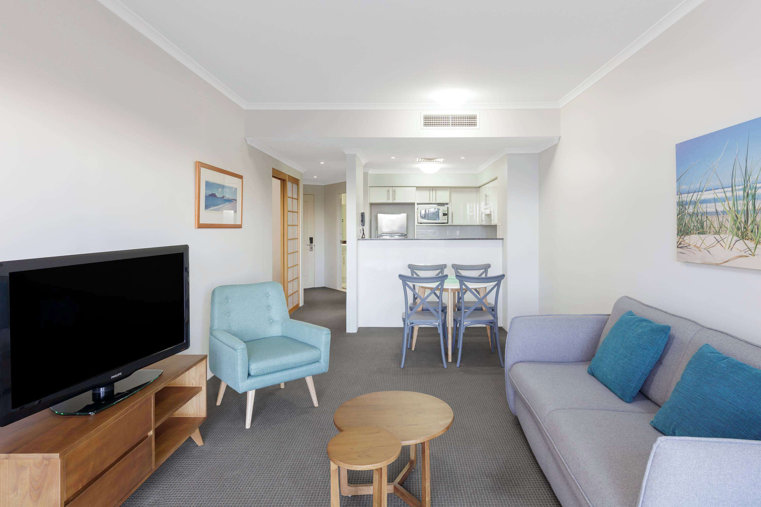 A living room with a flat-screen TV, a blue armchair, a grey sofa with blue cushions, a small wooden table, and a dining area with chairs and a microwave.