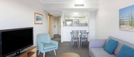 A living room with a flat-screen TV, a blue armchair, a grey sofa with blue cushions, a small wooden table, and a dining area with chairs and a microwave.