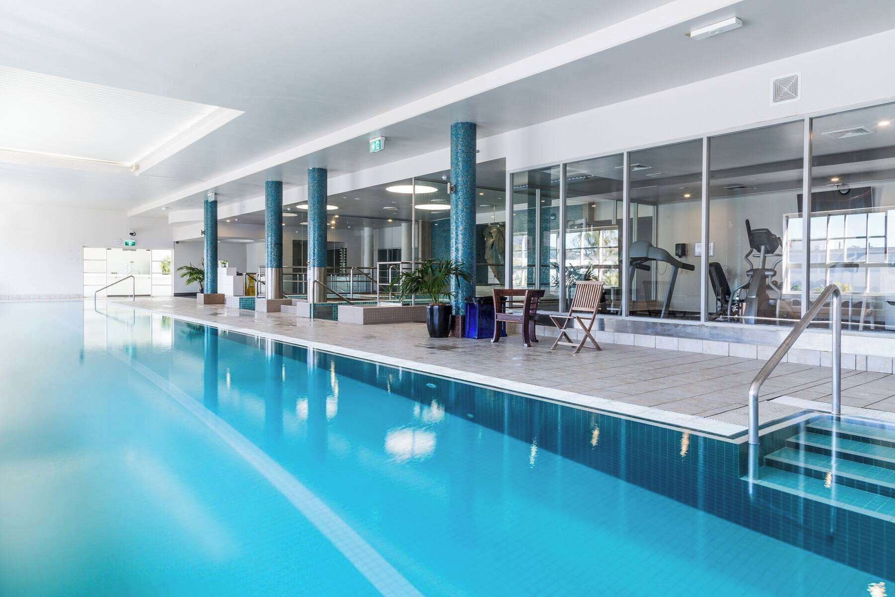 An indoor swimming pool with clear blue water, surrounded by glass walls and blue-tiled pillars.