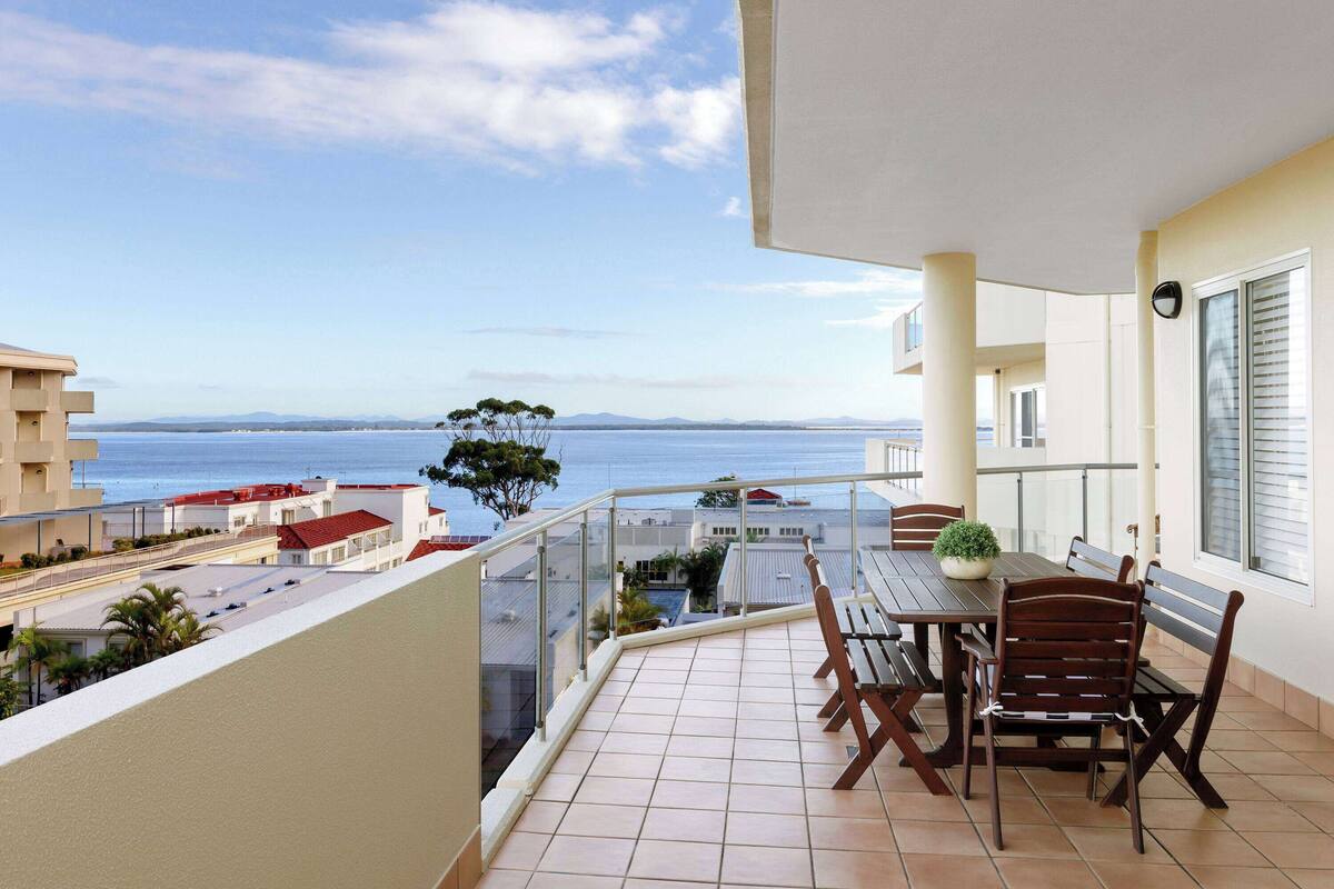 Ein Balkon mit Tisch und Stühlen, der Blick auf die Küste mit Gebäuden und Bäumen bietet.