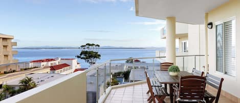 Un balcón con mesa y sillas, con vistas a la costa, edificios y árboles.