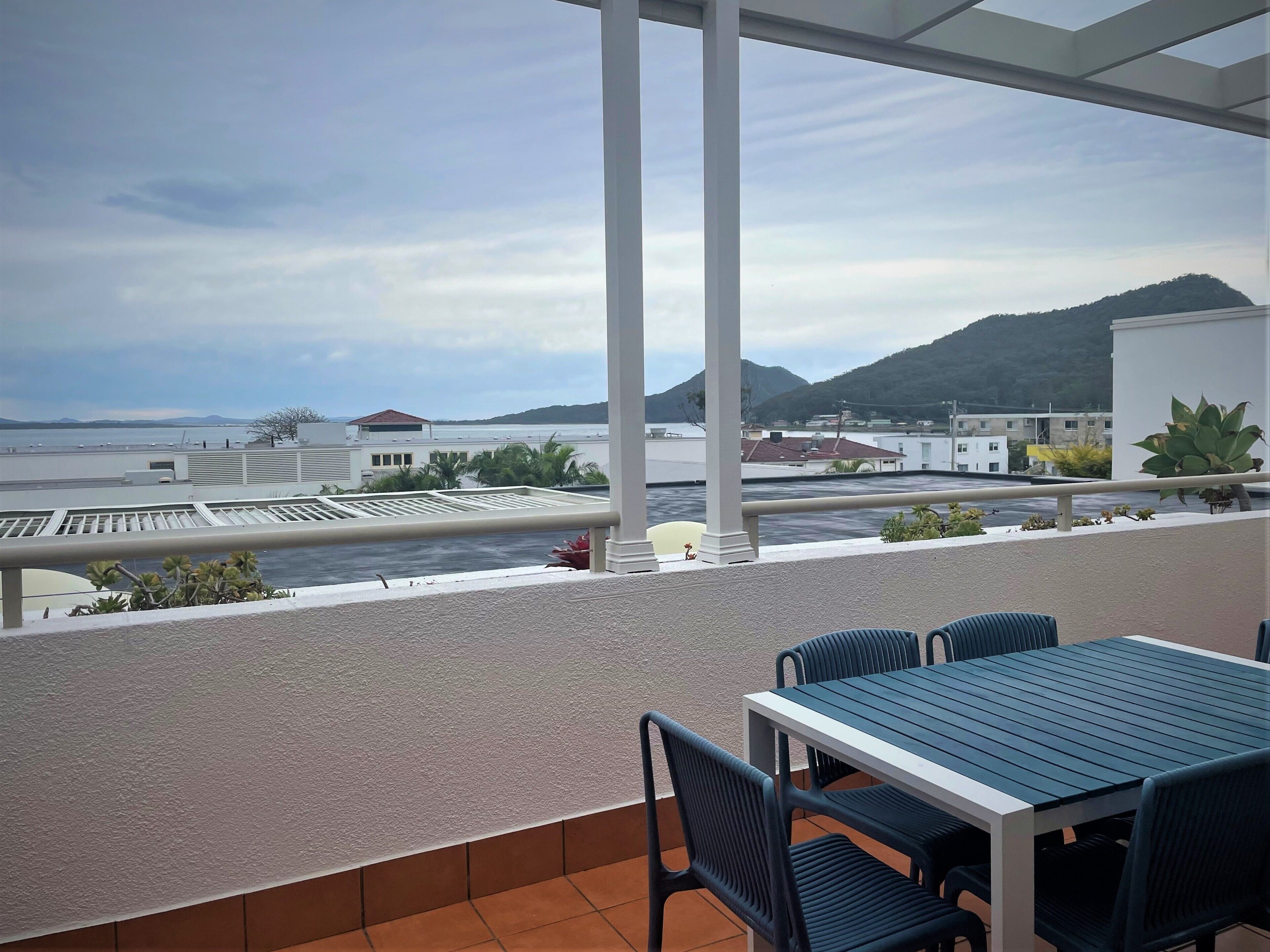 A balcony with a table and chairs, overlooking a coastal area with buildings and mountains.