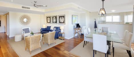 A modern living room with a glass dining table, white chairs, a blue sofa, and a ceiling fan.