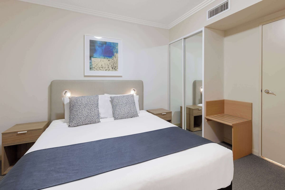 Ein Hotelzimmer mit einem großen Bett, Nachttischen, einem Spiegel, einer hölzernen Bank und einem gerahmten Kunstwerk an der Wand.