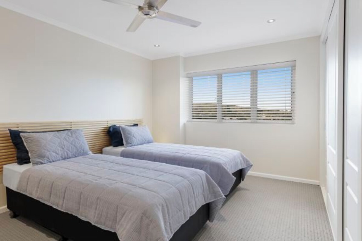 A bedroom with two beds, a ceiling fan, a window with blinds, and a carpeted floor.
