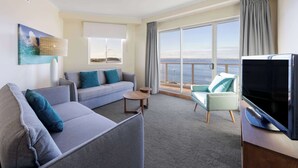 Een moderne hotelkamer met een grijze bank, een klein rond tafeltje, een televisie en uitzicht op de oceaan door grote ramen.