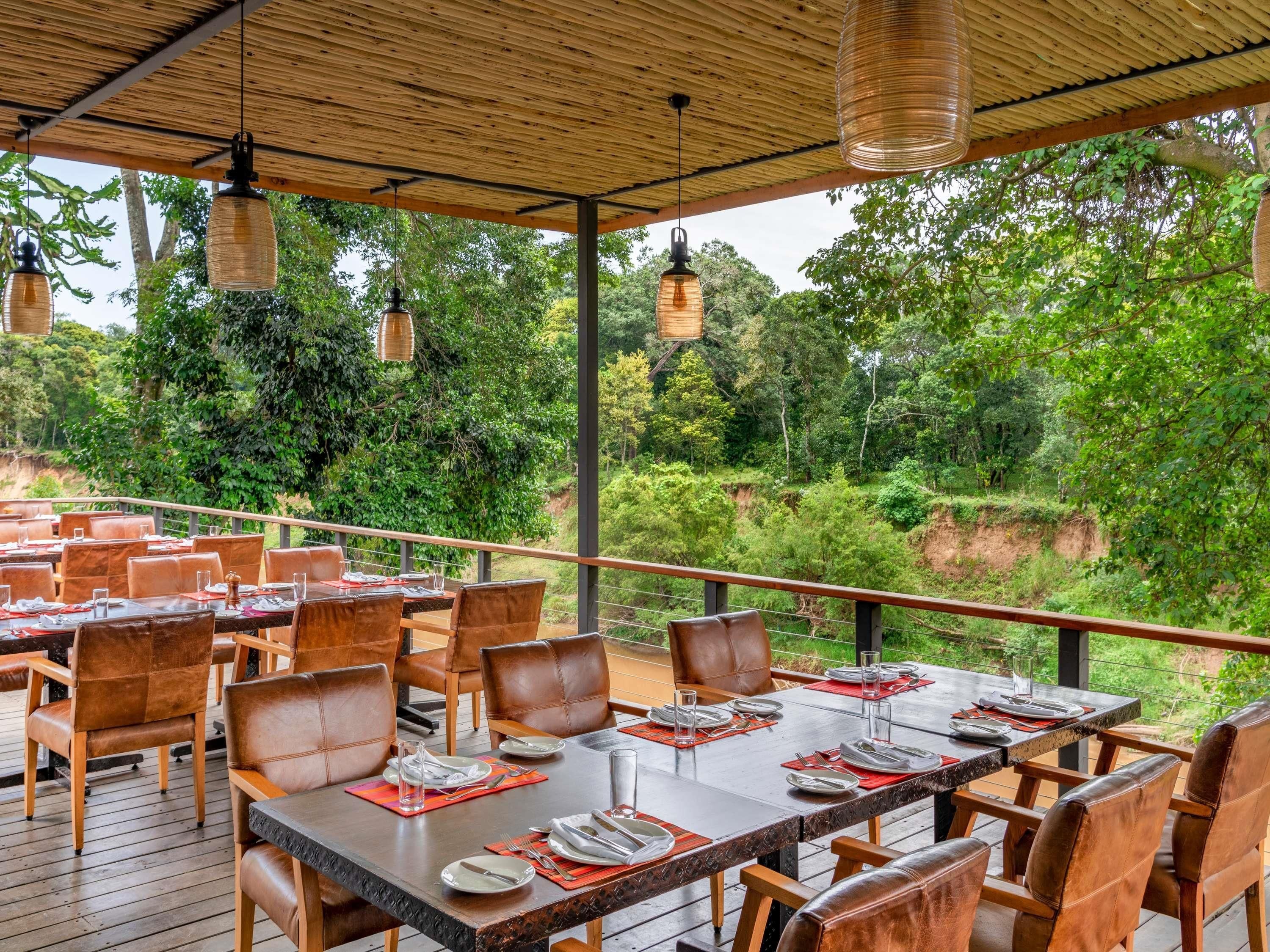 Un espacio de comedor al aire libre con mesas y sillas, rodeado de abundante vegetación y una terraza de madera.