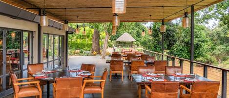An outdoor dining area with wooden tables and chairs, large windows, and hanging pendant lights.