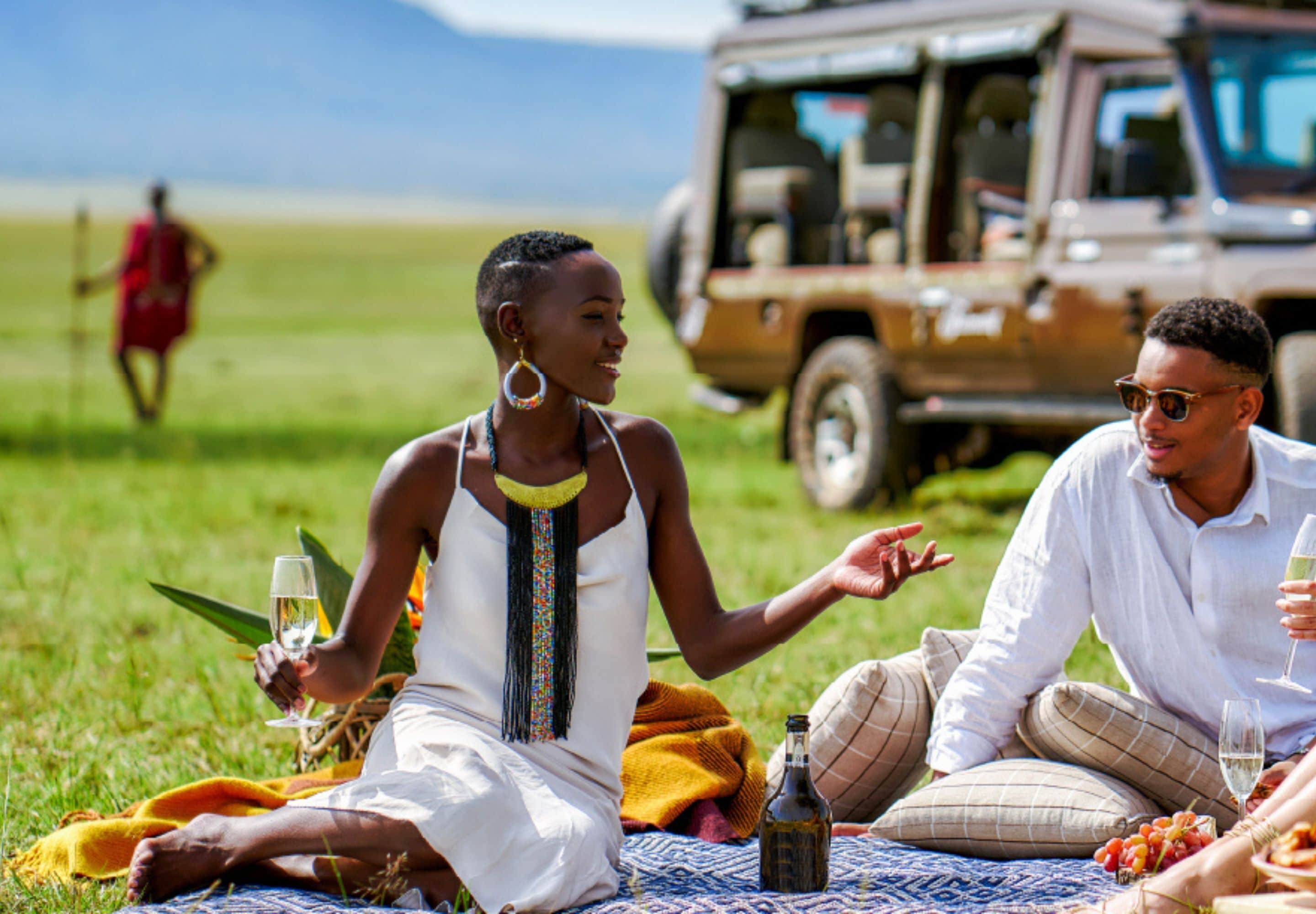 A group of people sitting on a blanket outdoors with a vehicle in the background.