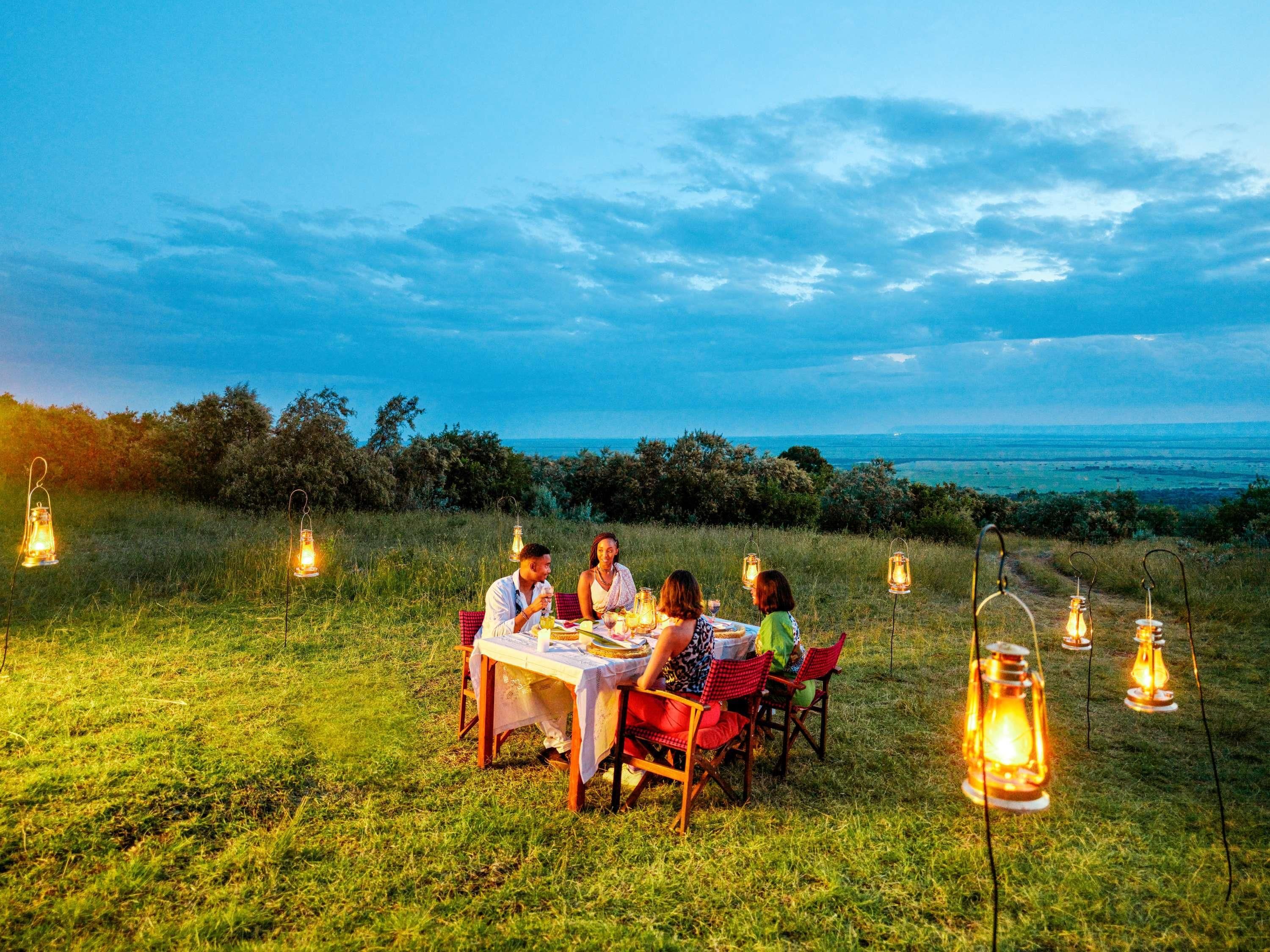 Um grupo de pessoas jantando ao ar livre, com uma mesa posta para refeição, rodeada por lanternas e uma vista panorâmica.