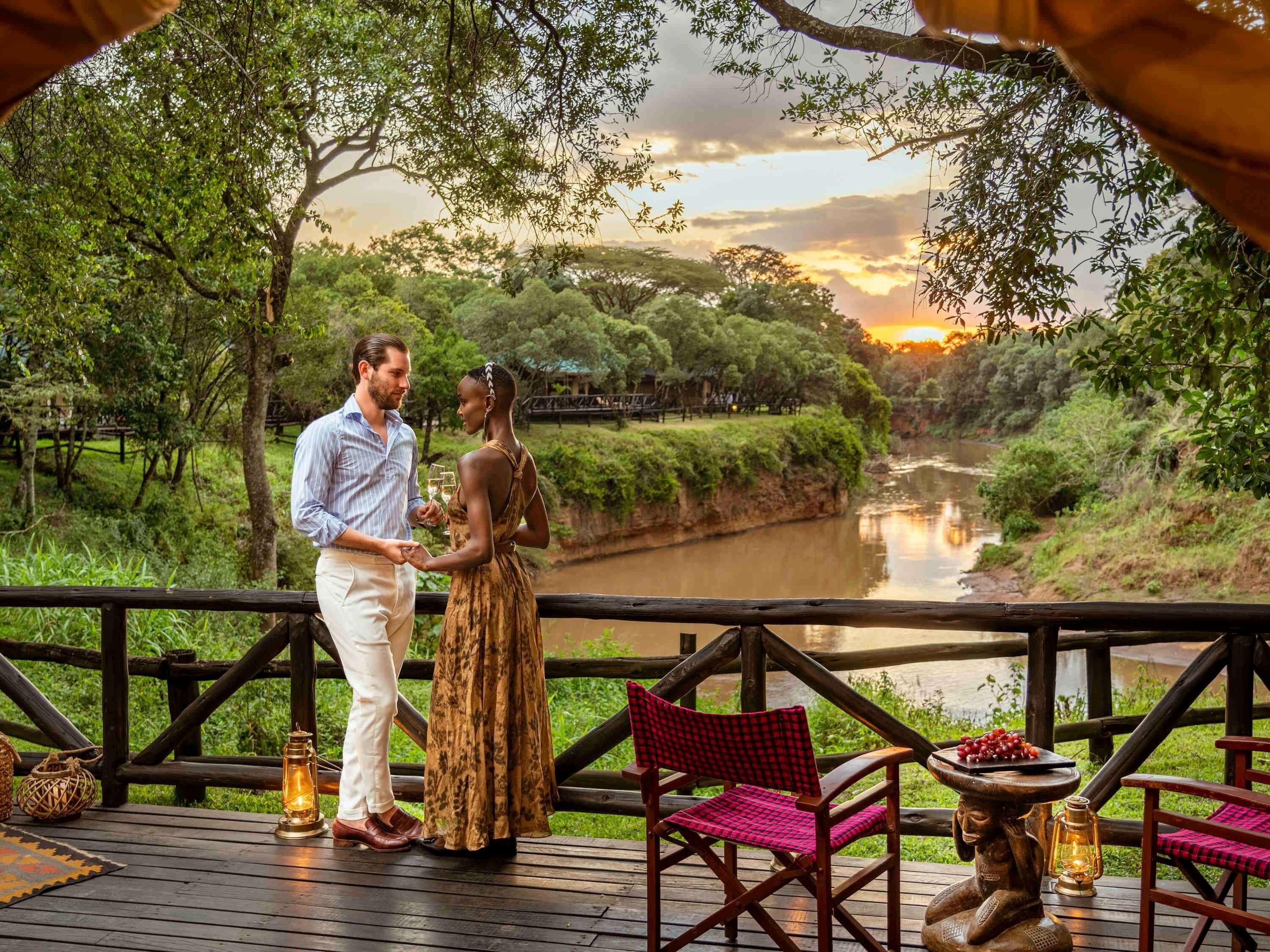 A couple standing on a wooden deck overlooking a river, with a sunset in the background.