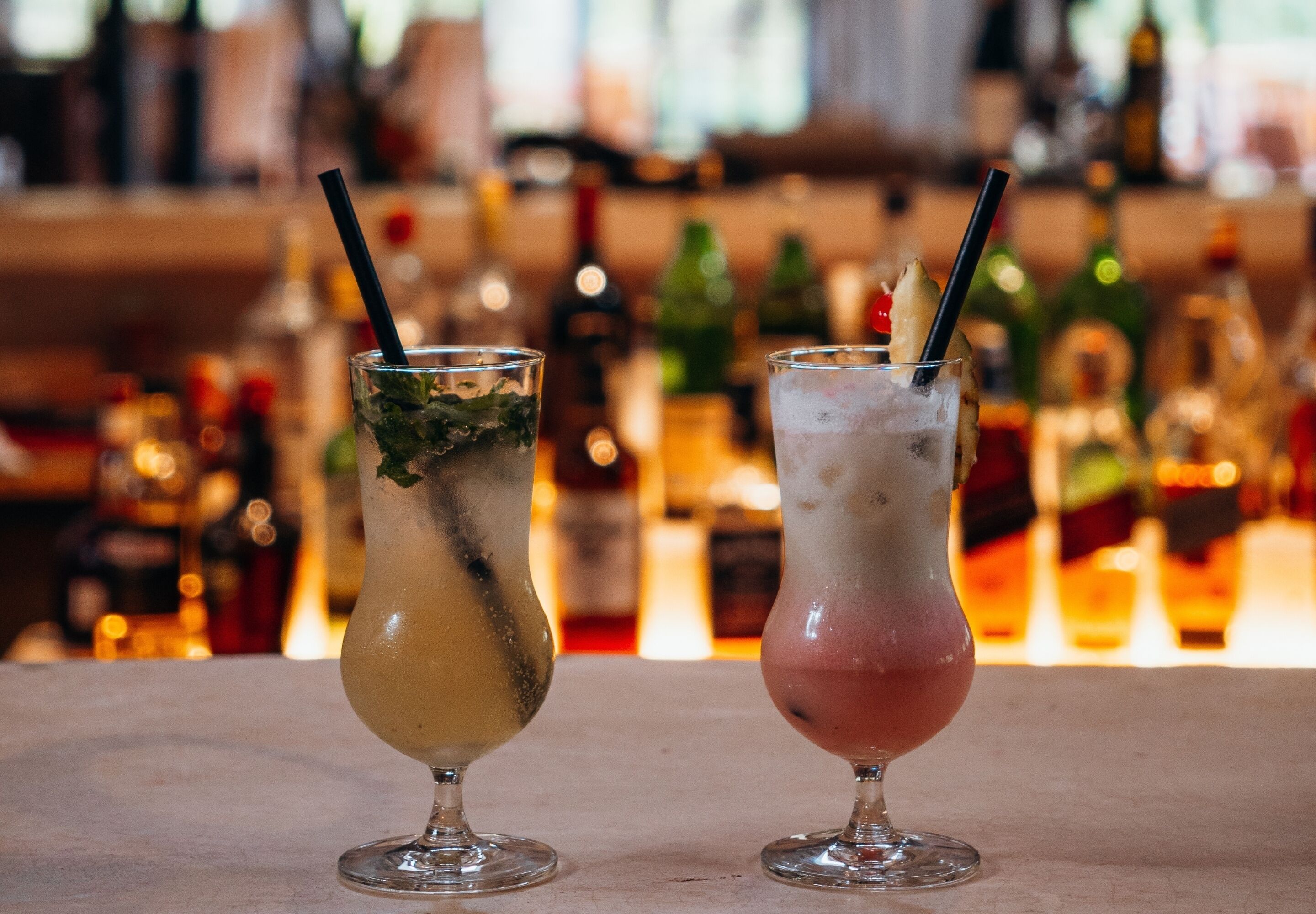 Zwei Cocktails in Longdrinkgläsern mit Strohhalmen, auf einer Bartheke mit Flaschen im Hintergrund.
