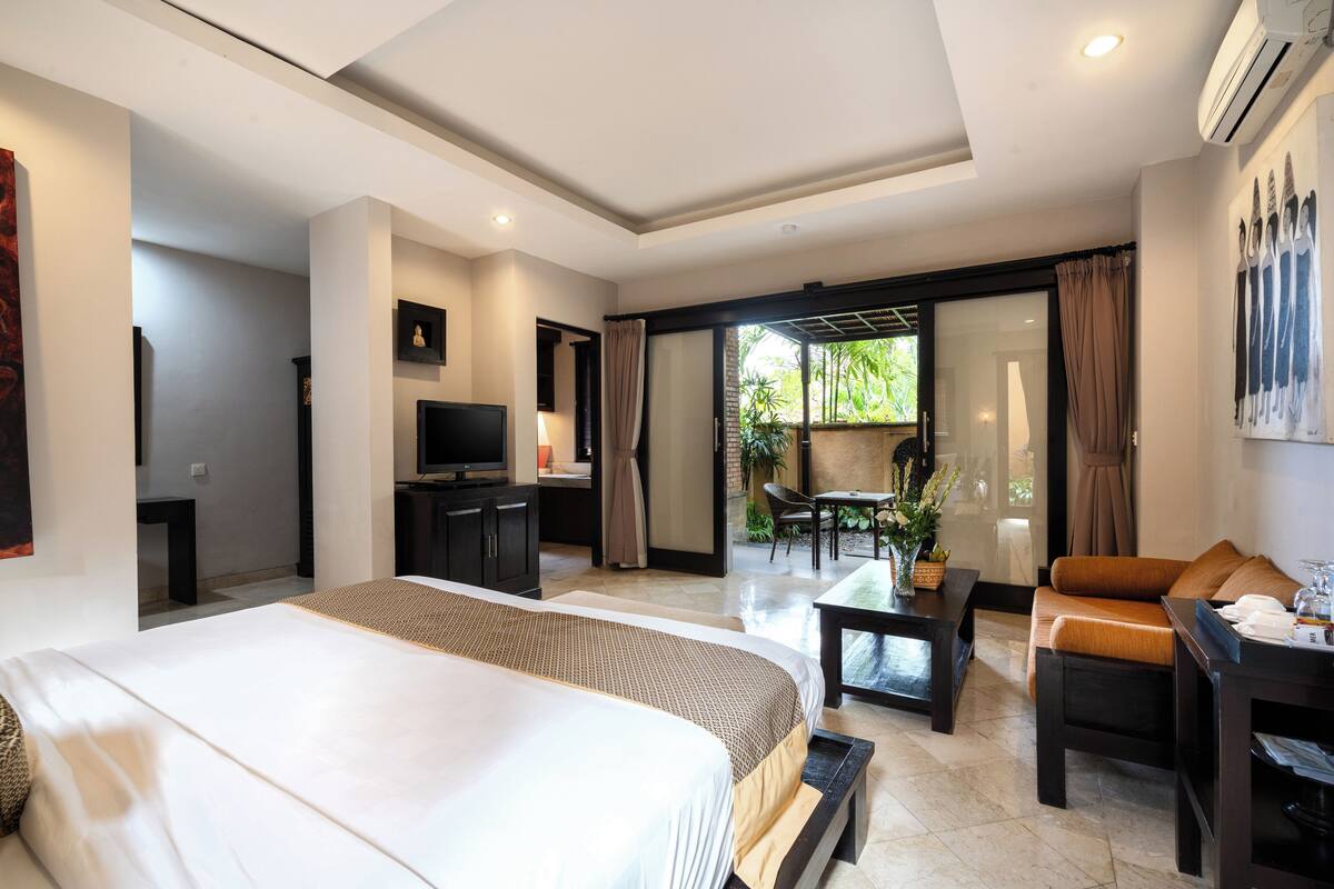 Ein Hotelzimmer mit einem großen Bett, einem Schreibtisch, einem Sessel, einem Fernseher und einer Glastür zum Balkon mit Pflanzen.