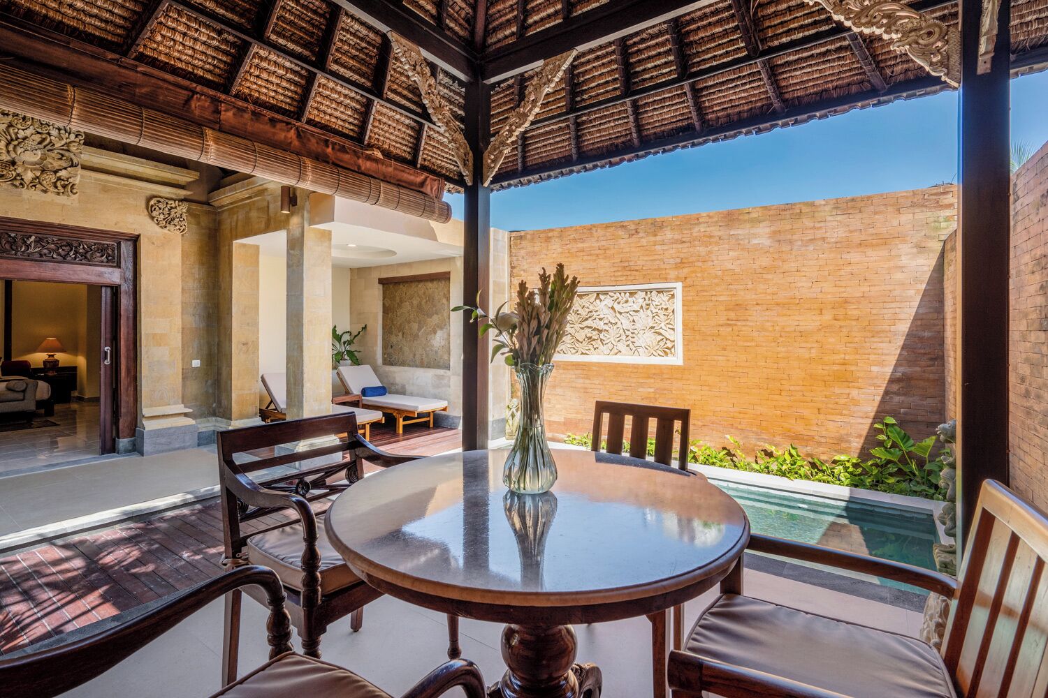 An outdoor seating area with a round table and chairs, a vase with flowers, and a view of a swimming pool and brick wall.