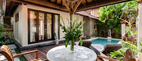 A wooden deck with a round table and chairs, a glass vase with plants, and a pool in the background.
