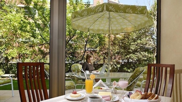 Mesa de desayuno al aire libre, con sombrilla, sillas y una persona al fondo.