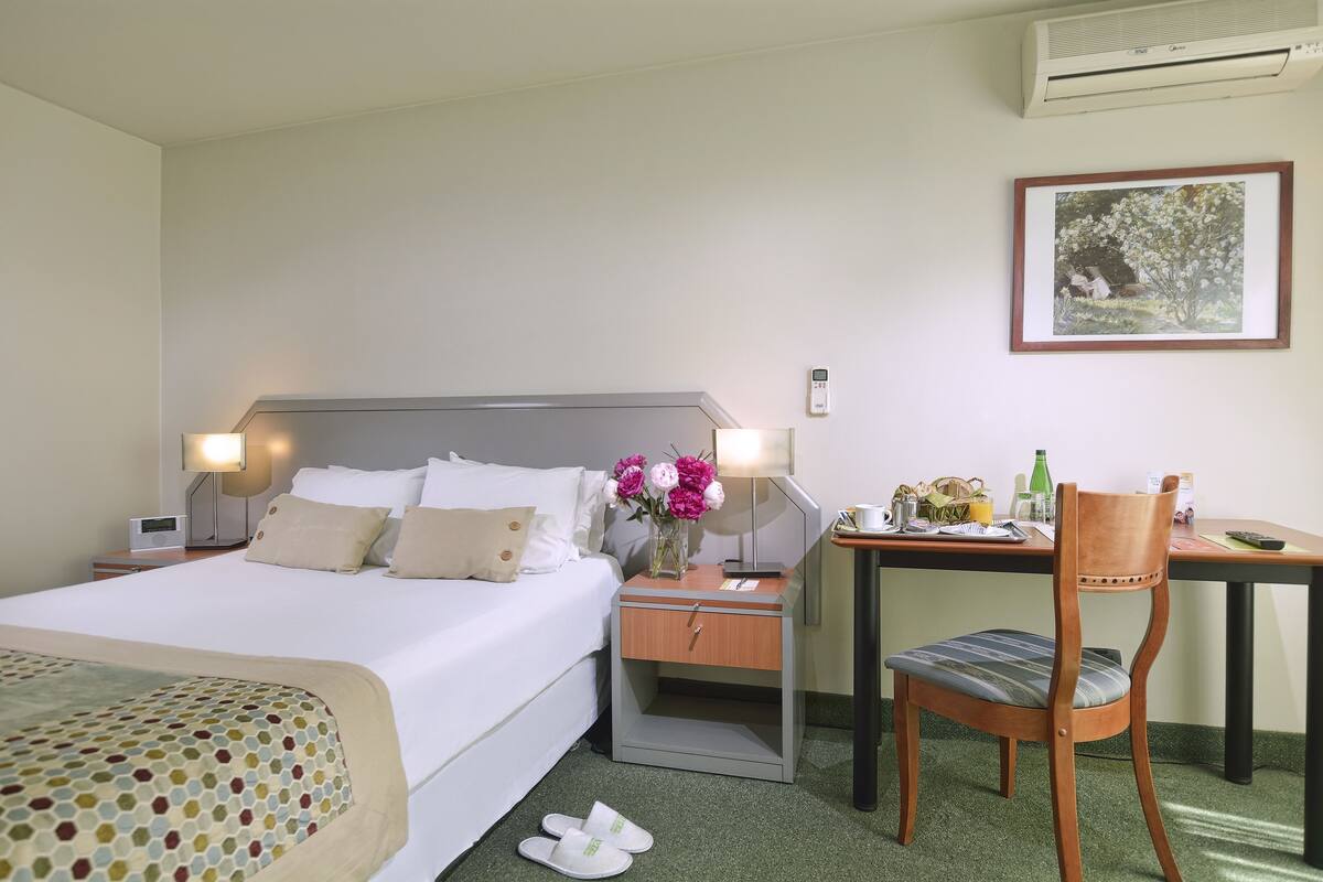 Habitación de hotel con cama, mesita de noche, silla, escritorio y un cuadro en la pared.