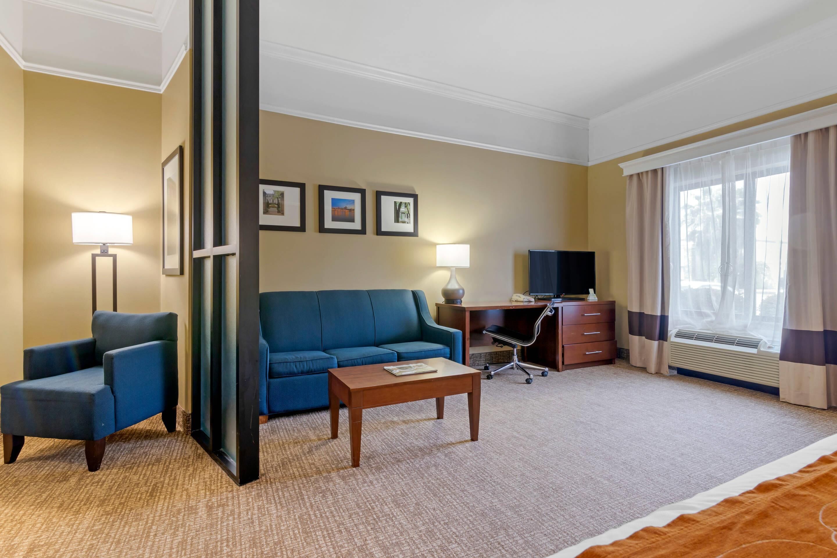 Ein Hotelzimmer mit blauem Sofa, einem Holztisch, einem Schreibtisch mit Stuhl, einem Flachbildfernseher und einem großen Fenster mit Vorhängen.