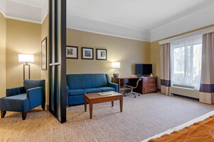 Habitación de hotel con un sofá azul, una mesa de centro de madera, un escritorio con una silla, un televisor de pantalla plana y un ventanal con cortinas.
