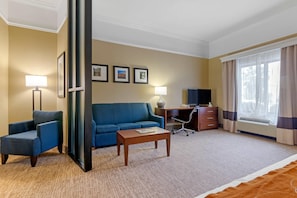 A hotel room with a blue sofa, a wooden coffee table, a desk with a chair, a flat-screen TV, and a large window with curtains.