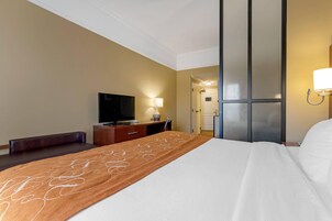 Quarto de hotel com uma cama, televisão, escrivaninha com luminária e porta para o banheiro.