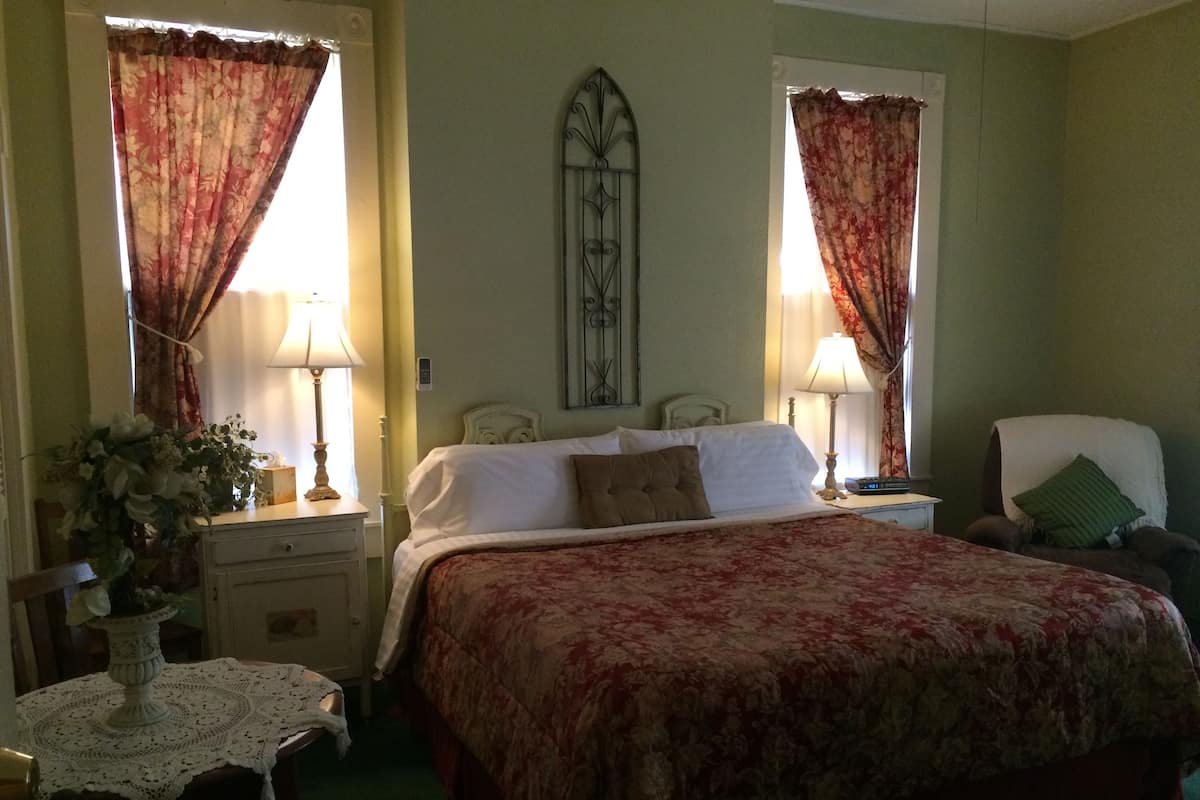 Une chambre à coucher comprenant un lit, deux tables de chevet avec des lampes, une chaise, une table d’appoint et des rideaux.