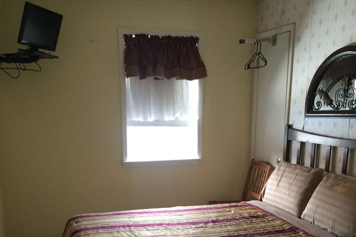 Une chambre à coucher avec un lit, une fenêtre avec des rideaux, un téléviseur fixé au mur et un porte-manteau.
