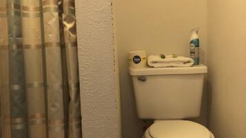 Un baño con inodoro, un rollo de papel higiénico, una botella de jabón para manos y una cortina de ducha.