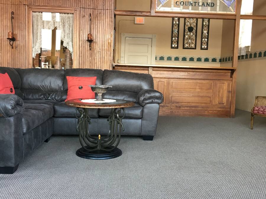 A cozy living room with a leather sectional sofa, a vintage table, and a ceiling fan. A wooden door with a sign reading "COURTLAND" is visible in the background.