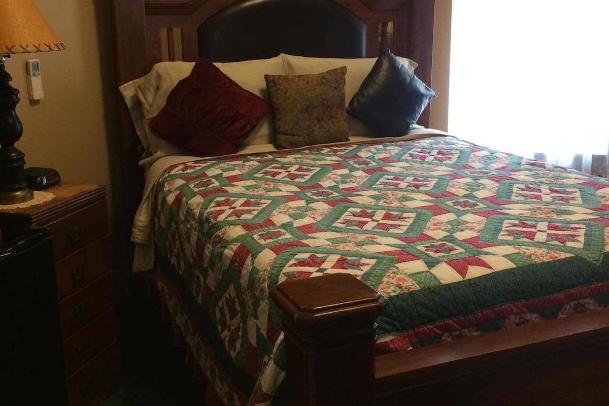 Un lit bien fait, avec une tête de lit à motifs floraux, une table de chevet en bois surmontée d’une lampe, et une fenêtre ornée de rideaux légers.