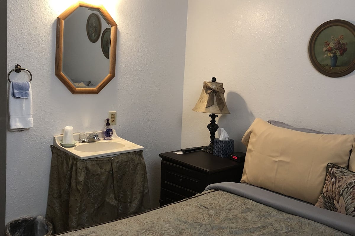 Une chambre d’hôtel comprenant un lit, une table de chevet avec une lampe, une petite commode, un miroir, un tableau et une salle de bain avec un lavabo et un porte-serviettes.
