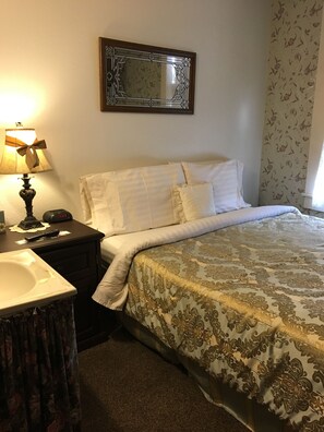 Habitación de hotel con una cama, una mesita de noche, una lámpara, un espejo y una ventana con cortinas florales.