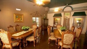 Comedor con una gran mesa de madera y sillas ornamentadas, ventilador de techo y obras de arte enmarcadas en las paredes.