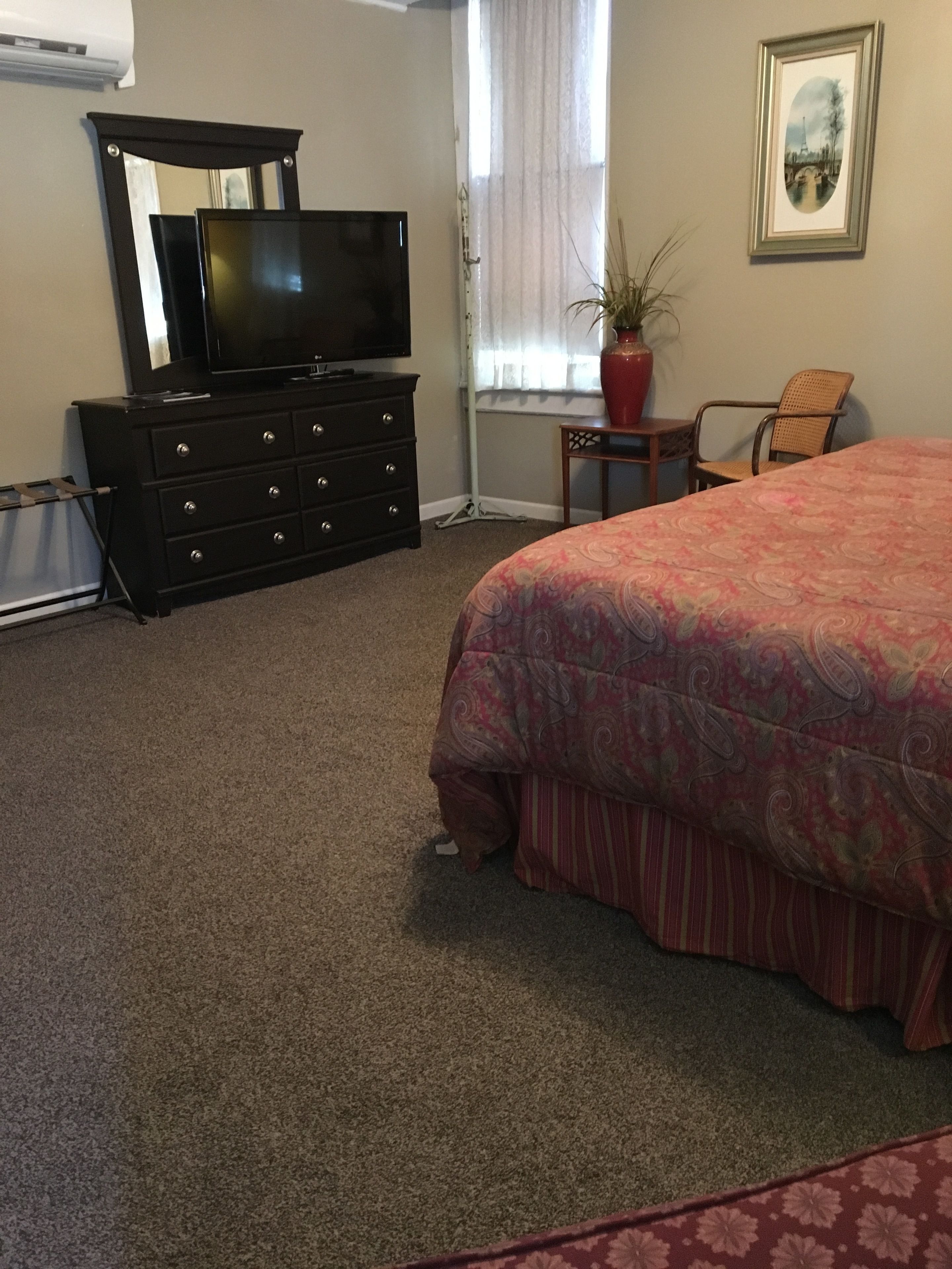Un dormitorio con cama, cómoda, televisor, silla y ventana con cortinas.