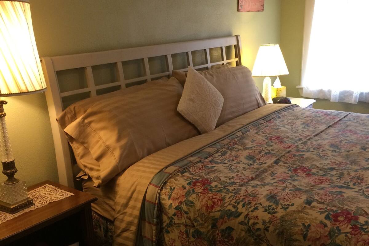 Un lit bien fait, recouvert d’une couvre-lit à motifs floraux, une lampe de chevet et une petite table de chevet sur laquelle se trouvent une lampe et un objet décoratif.