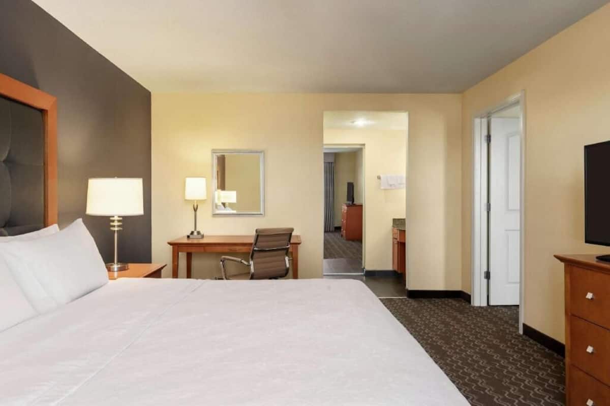A hotel room with a bed, desk, chair, lamp, television, and a door leading to another room.