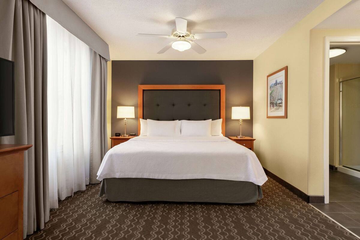 A hotel room with a bed, two bedside lamps, a ceiling fan, a framed picture on the wall, and a door leading to another room.