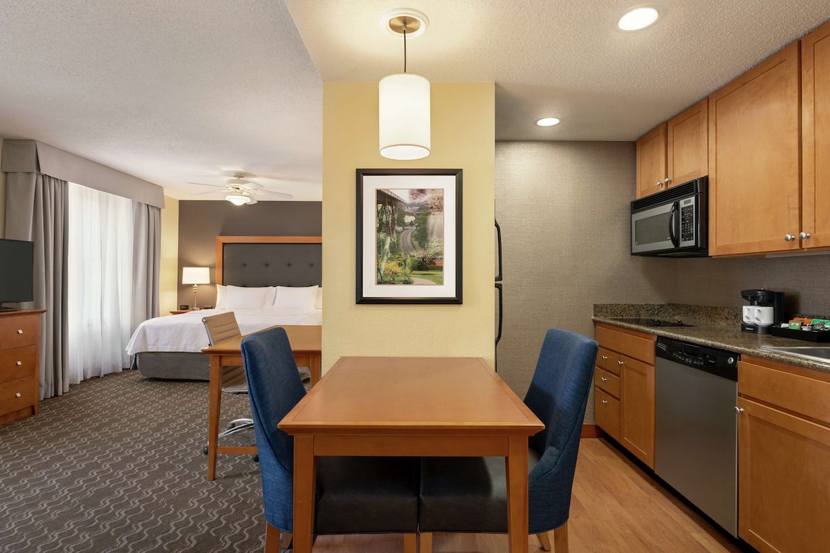 A hotel room with a bed, a desk with chairs, a kitchenette, and a framed picture on the wall.