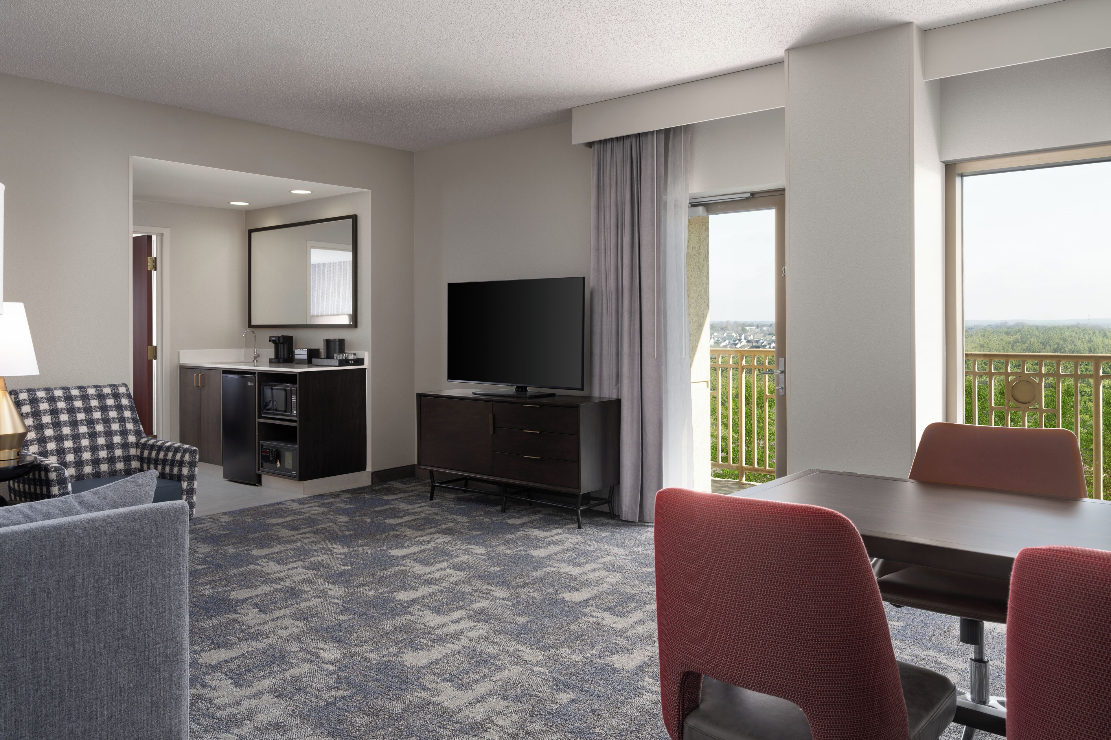 Habitación de hotel con televisión de pantalla plana, una mesa de comedor con sillas, un sofá, una pequeña cocina y un balcón con vistas.