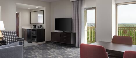 Habitación de hotel con televisión de pantalla plana, una mesa de comedor con sillas, un sofá, una pequeña cocina y un balcón con vistas.