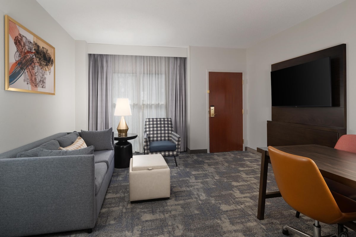 Une chambre d’hôtel équipée d’un canapé, d’un pouf, d’un fauteuil, d’une table, d’une télévision et de rideaux.