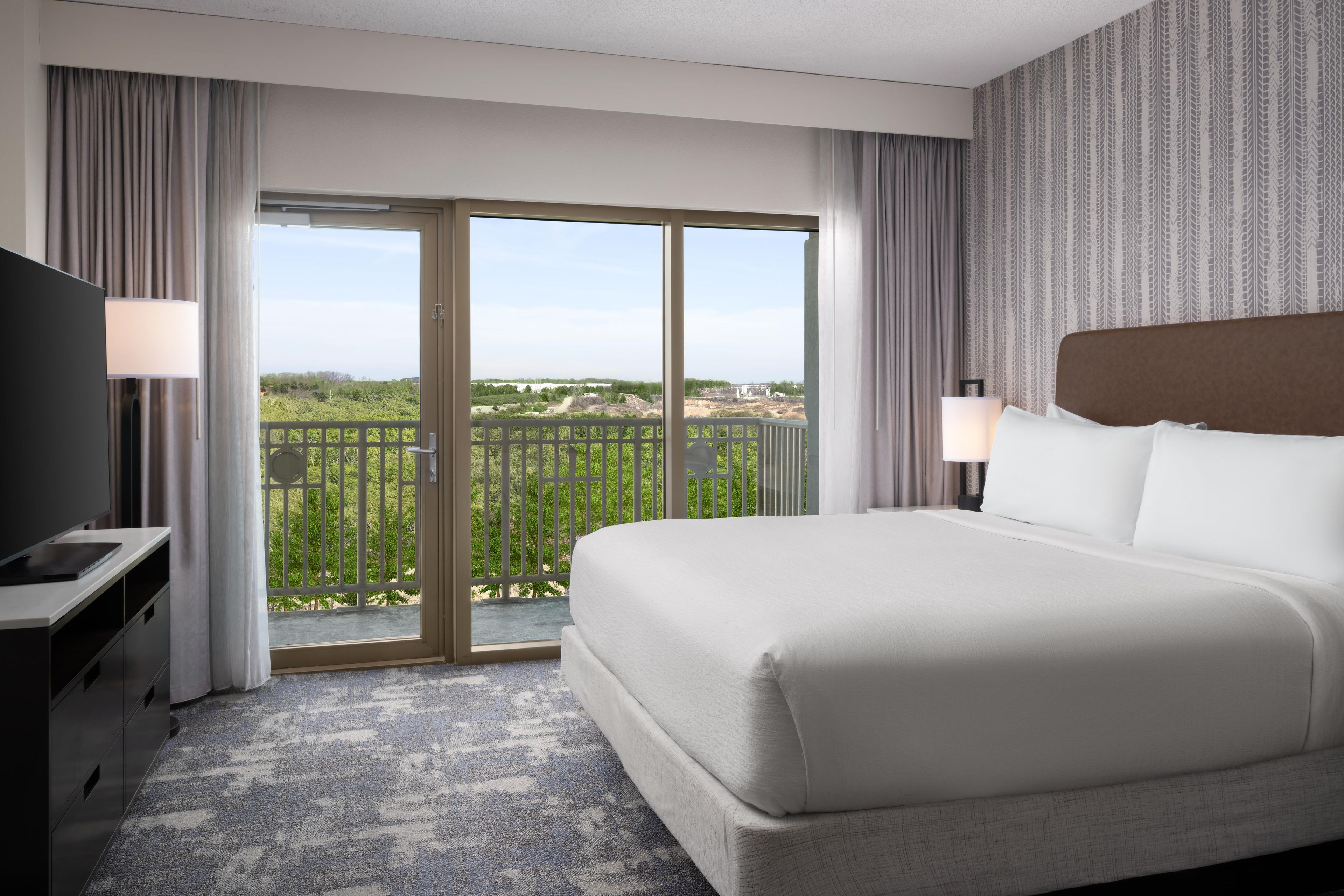 A hotel room with a large bed, a TV, a desk, and a balcony with a view.
