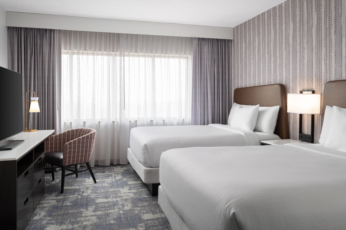 Ein Hotelzimmer mit zwei Betten, einem Schreibtisch, einem Sessel und einem Fernseher.