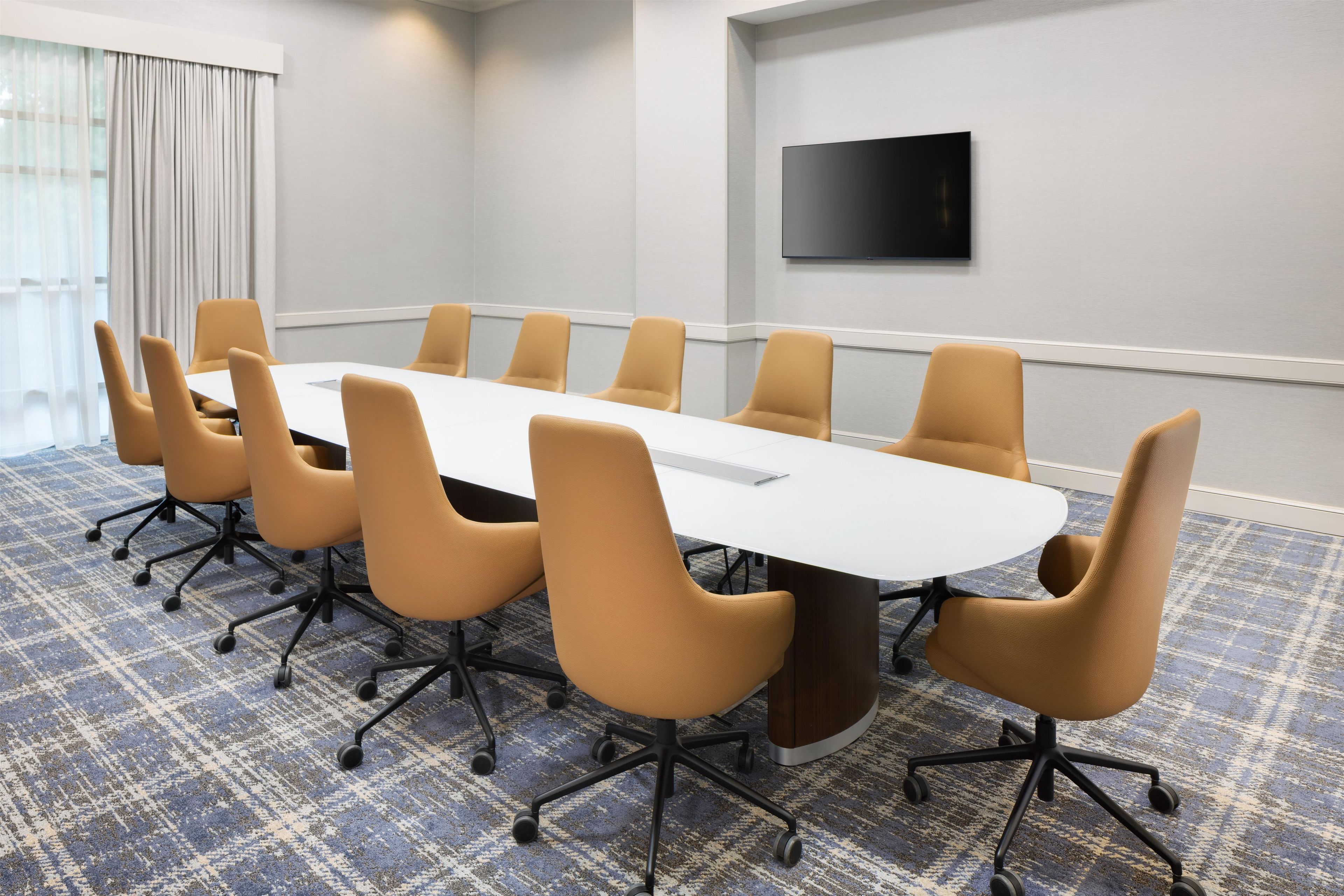 Een vergaderruimte met een lange tafel, zes stoelen, een flatscreen televisie aan de muur en vloerbedekking met een patroon.