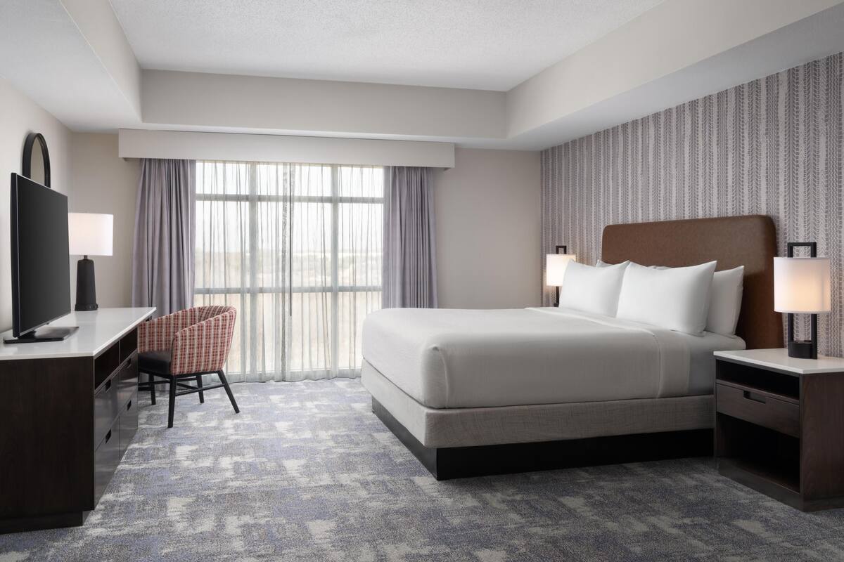 Une chambre d’hôtel avec un lit, un bureau, une chaise, une télévision et une fenêtre avec des rideaux.