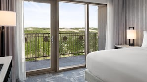 Habitación de hotel con una cama grande, un escritorio, una silla, un balcón con vistas a la vegetación y una ventana con cortinas.