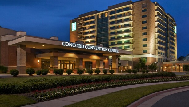 Un hotel de varios pisos con un llamativo letrero que dice 'CONCORD CONVENTION CENTER', una entrada ajardinada y un diseño arquitectónico moderno.
